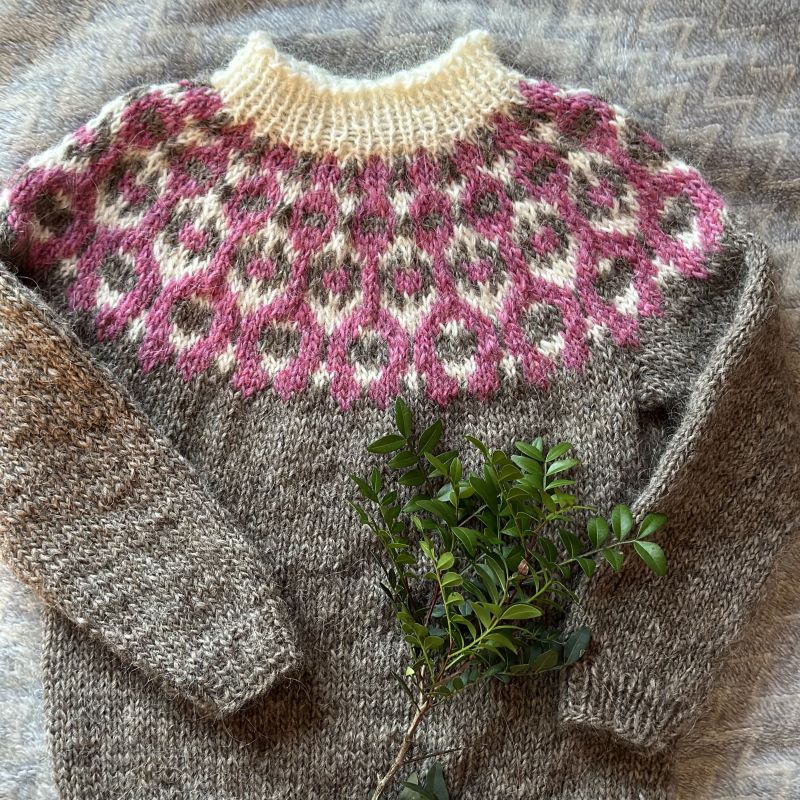 A grey, pink and white hand knitted children's Icelandic lopapeysa sweater with a geometric pattern and a round neckline, on a wooden surface.