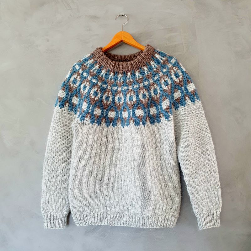 A gray, handknitted Icelandic lopapeysa sweater with a brown and blue geometric pattern, including a round neckline and long sleeves, shown on a hanger.