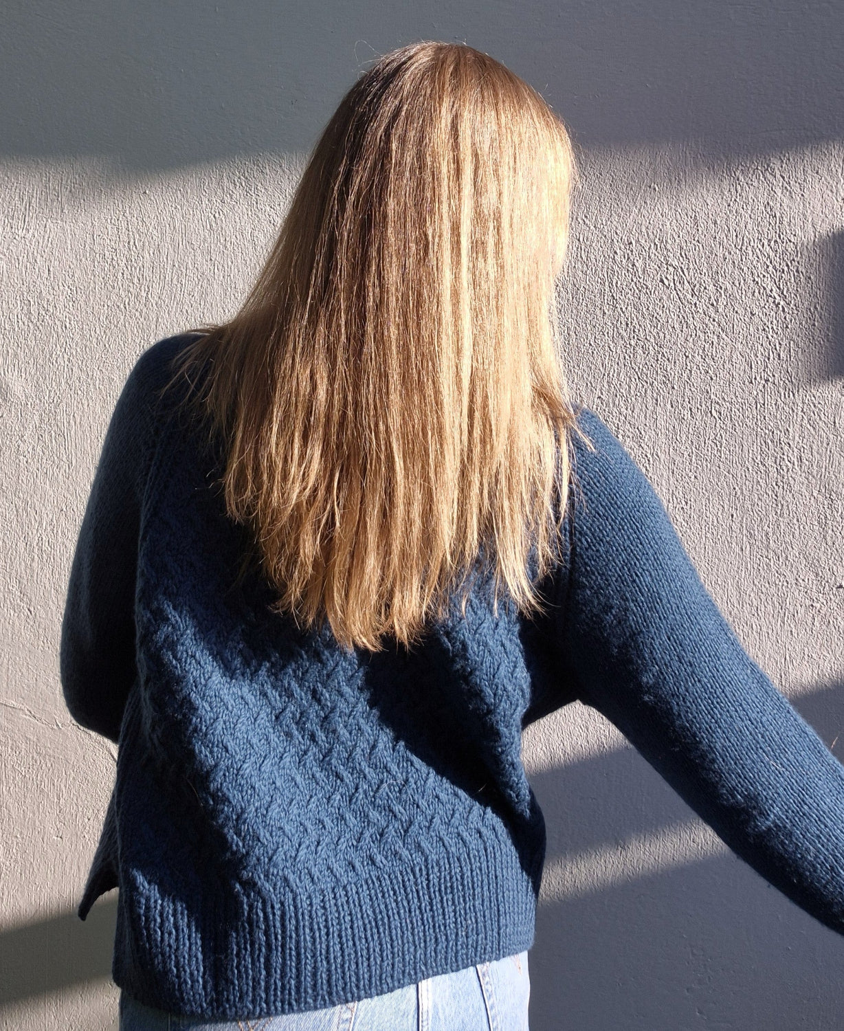 Woman wearing a beautiful blue  hand knitted sweater with textured pattern, against a plain background, seen from behind.