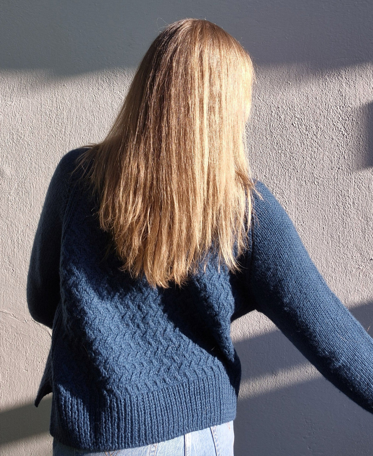 Woman wearing a beautiful blue  hand knitted sweater with textured pattern, against a plain background, seen from behind.