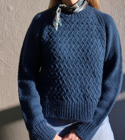 Woman wearing a beautiful blue  hand knitted sweater with textured pattern, against a plain background.