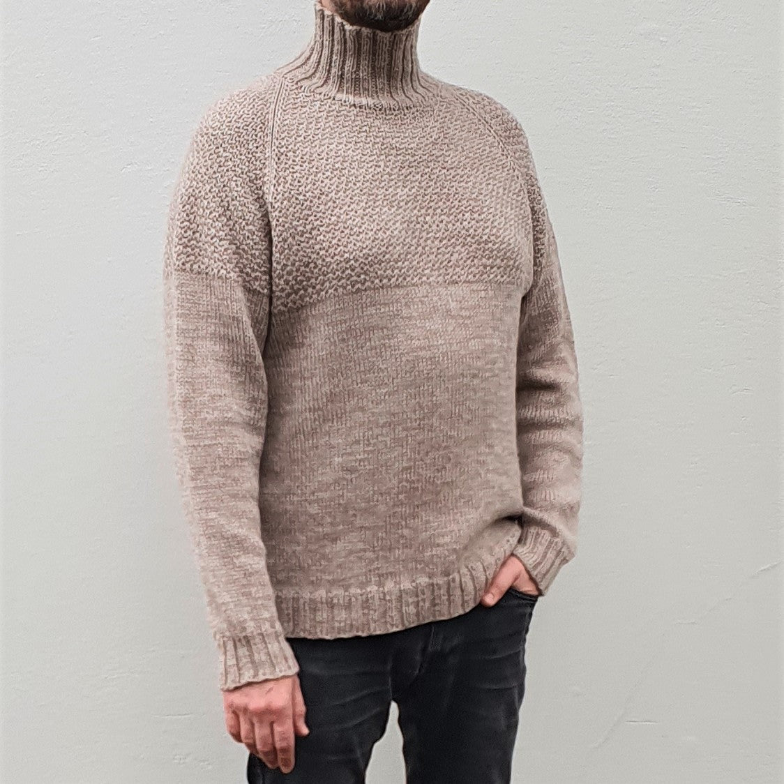 A man wearing a beige, high-collared, long-sleeved Duggari sweater, standing against a neutral background.