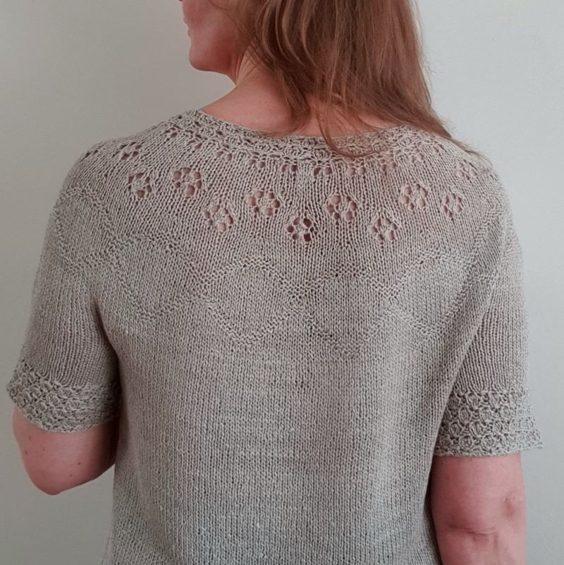 A woman wearing a short-sleeved, knitted top with a round neckline and decorative eyelets on the yoke, seen from behind.