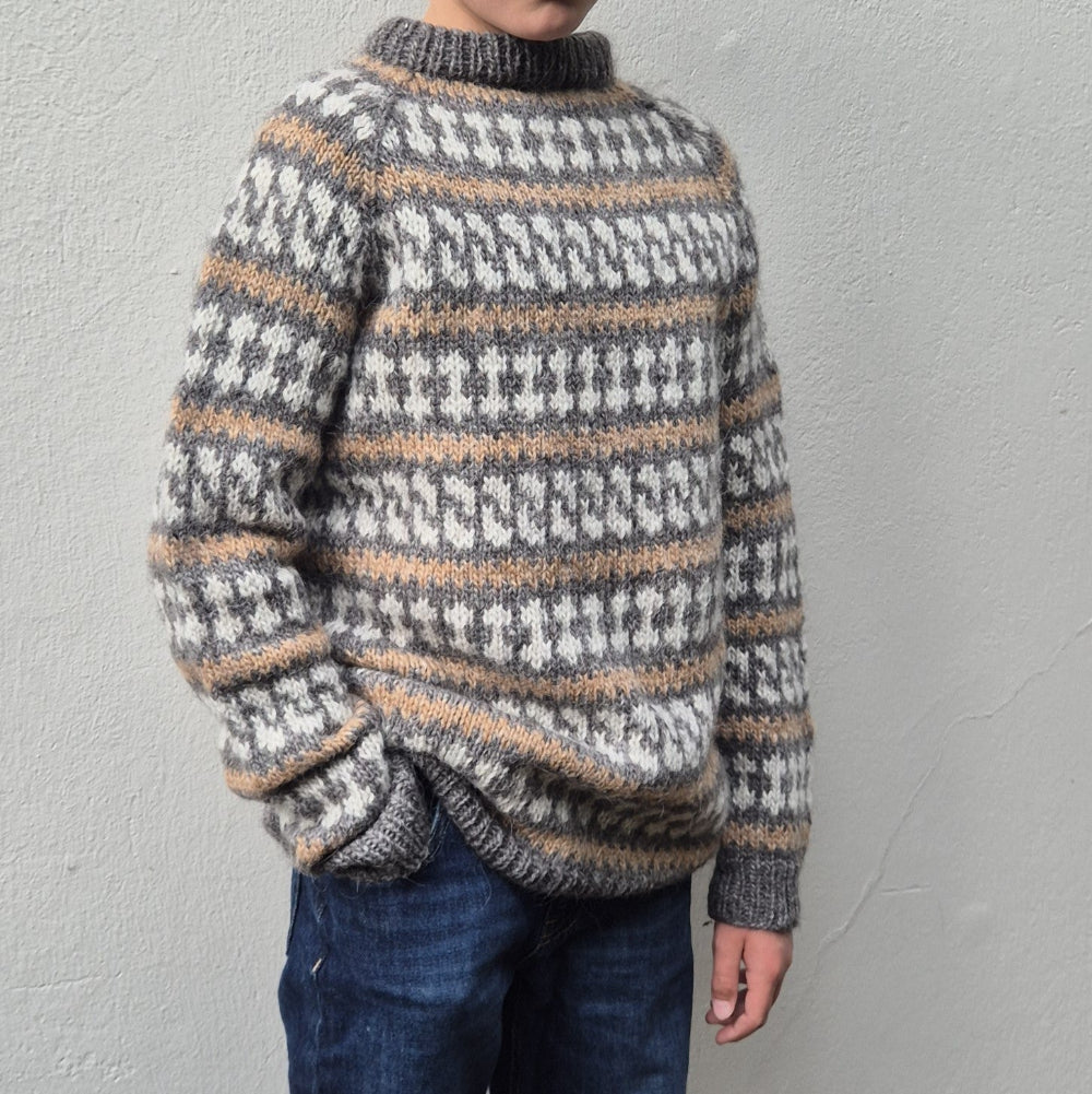 A kid wearing a handknit Nordic style Freyr lopapeysa sweater with a gray, white, and tan Fair Isle pattern.