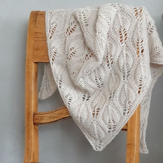 An exclusive light beige cashmere hand knitted scarf with a flowing leaf pattern, draped over the back of a wooden chair.