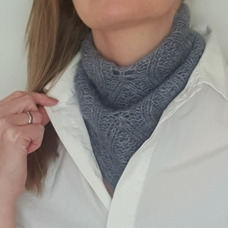 A woman wearing a small blue refined  hand knitted scarf with a flowing leaf pattern, styled with a minimalistic white shirt.