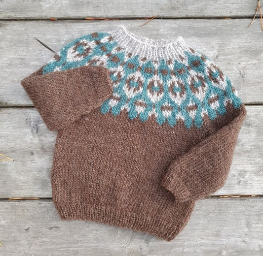 A brown and beige colored hand knitted children's Icelandic lopapeysa sweater with a geometric pattern and a round neckline, on a wooden surface.