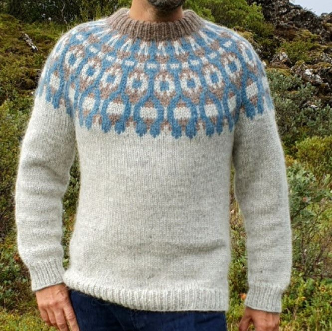 A person standing outdoors wearing a gray, handknitted Icelandic lopapeysa sweater with a brown and blue geometric pattern, including a round neckline and long sleeves.
