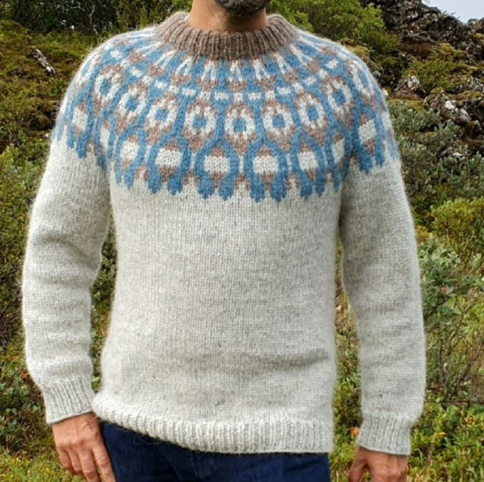 A person standing outdoors wearing a gray, handknitted Icelandic lopapeysa sweater with a brown and blue geometric pattern, including a round neckline and long sleeves.