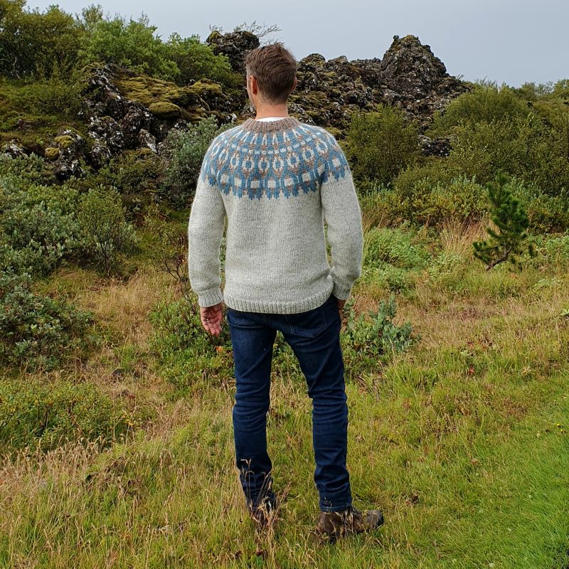 A person standing outdoors wearing a gray, handknitted Icelandic lopapeysa sweater with a brown and blue geometric pattern, including a round neckline and long sleeves.